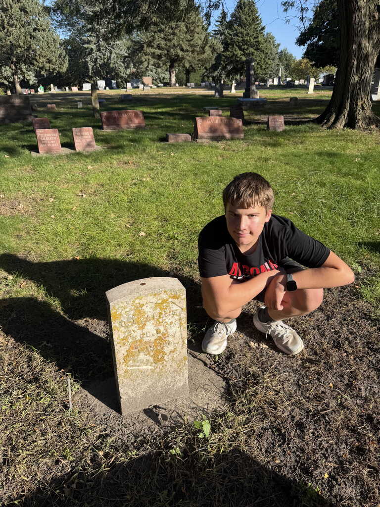 A person kneels next to a gravestone in a cemetery with green grass and trees.