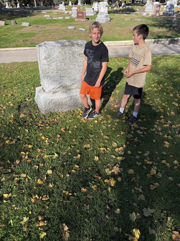Two children stand in a cemetery, with one boy by a large tombstone and the other boy nearby.