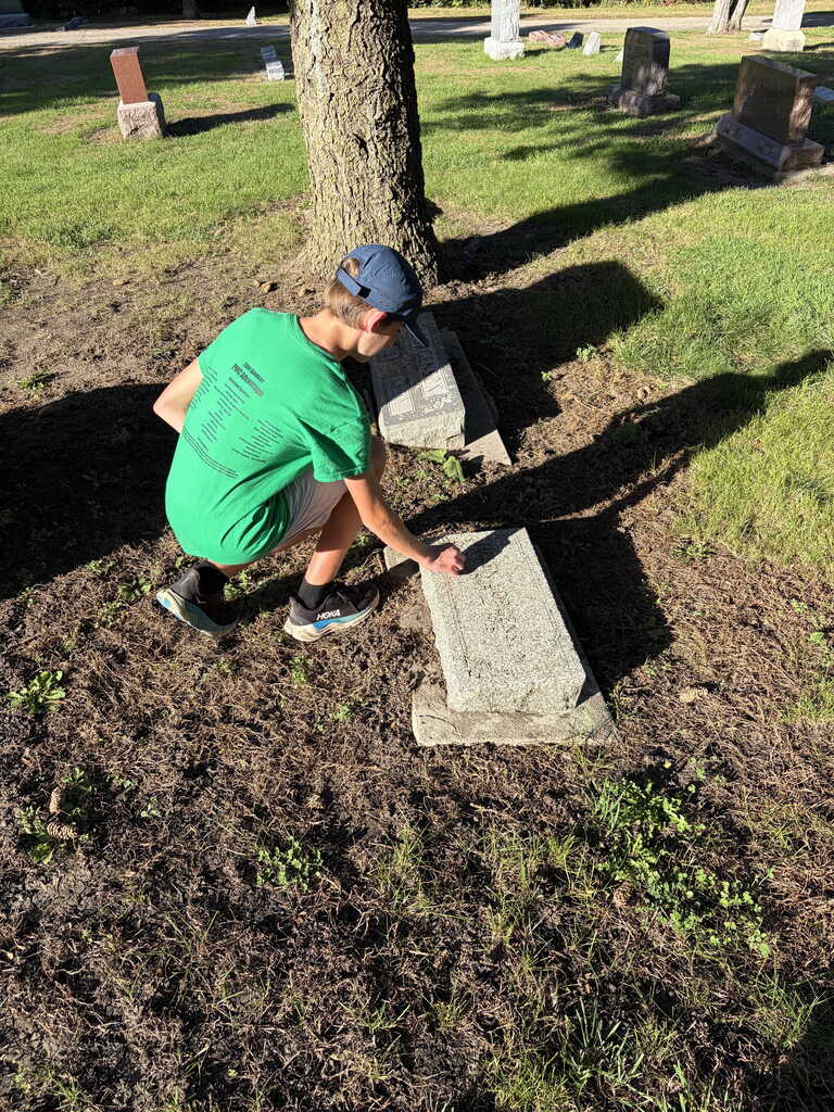 Person in a green shirt and blue hat cleaning a gravestone in a cemetery.