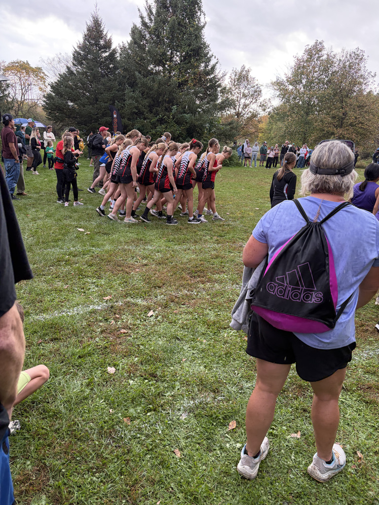 A group of runners in red and black uniforms warms up on a grassy field. Spectators watch. Trees and cloudy sky in background.