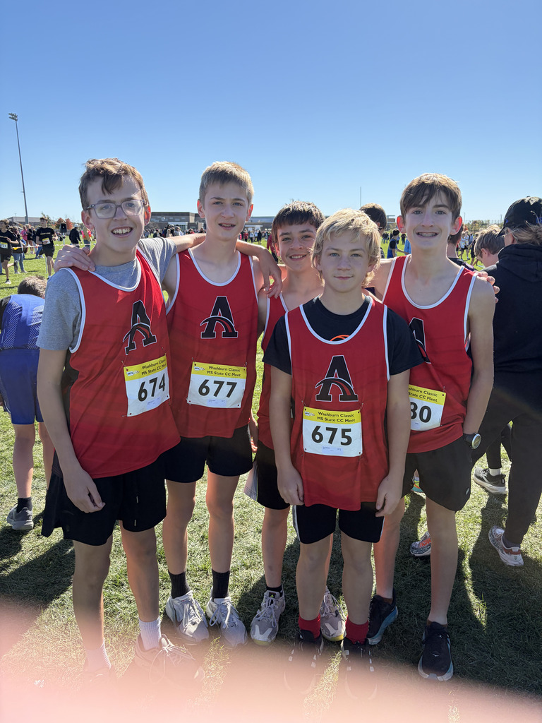 Five boys in matching red vests with numbers, standing in a line on a grassy field.