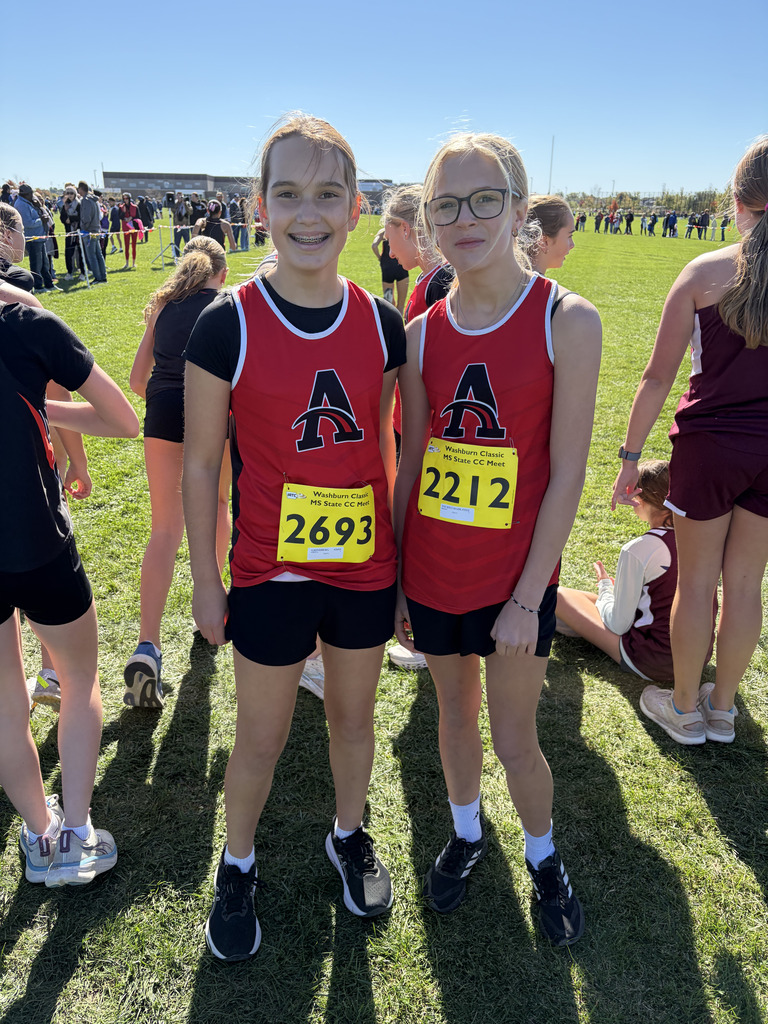 Two girls in red shirts with number tags stand on a grassy field with other people.