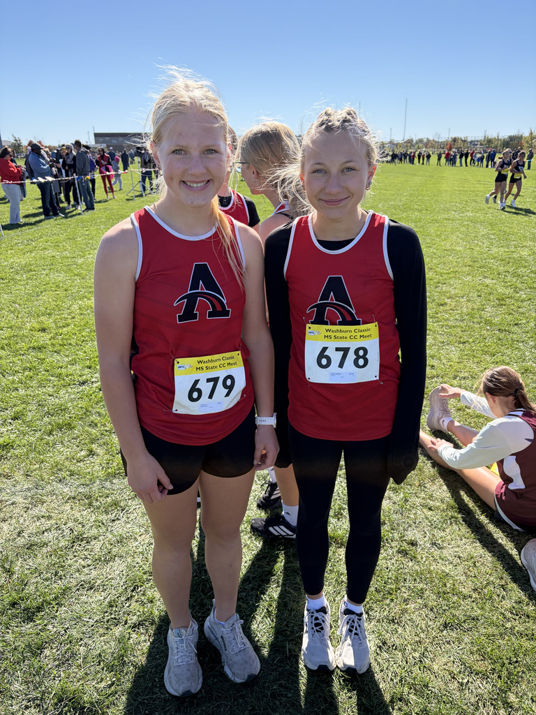 Two girls stand in a grassy field, both wearing red and black athletic uniforms with numbers.