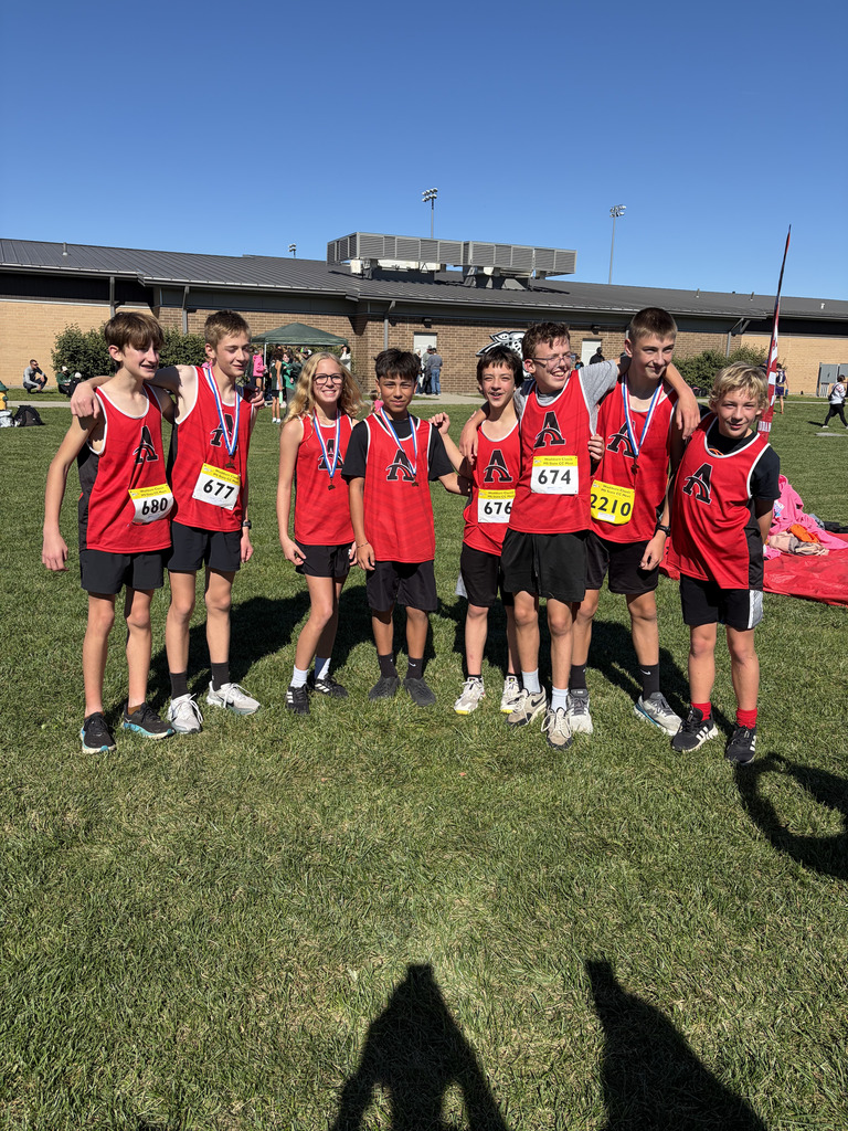 Group of seven young students in red shirts and medals stand on grass, school building in background.