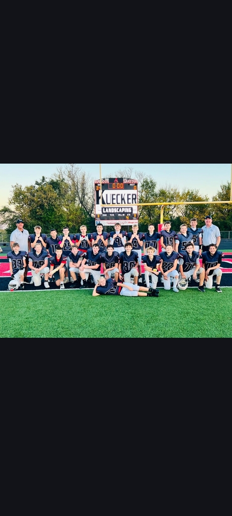 Football team poses on field with trees and large sign with white lettering in background.
