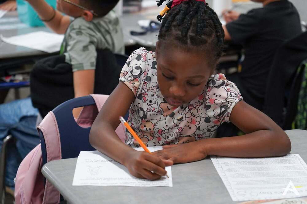 A student works on a written assignment at a classroom desk while holding a pencil. Other students are visible working in the background.