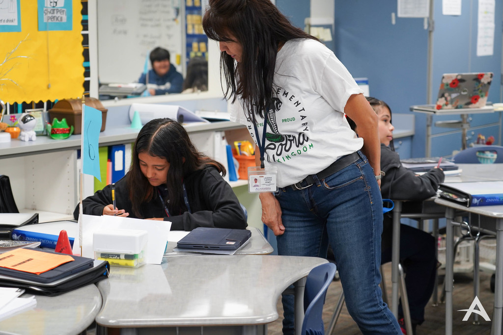 A teacher stands beside a student’s desk, reviewing the student’s work during a classroom activity.