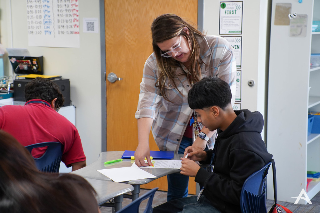 A teacher stands beside a seated student, pointing to a worksheet and offering guidance