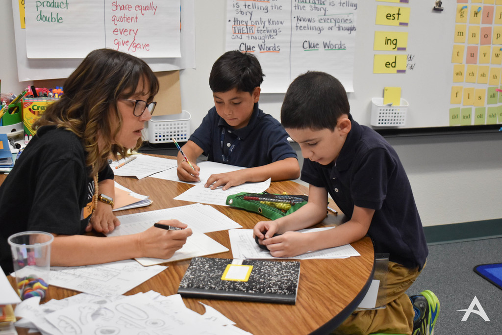 Teacher working one-on-one with two elementary students at a small table as they write on worksheets in a classroom.