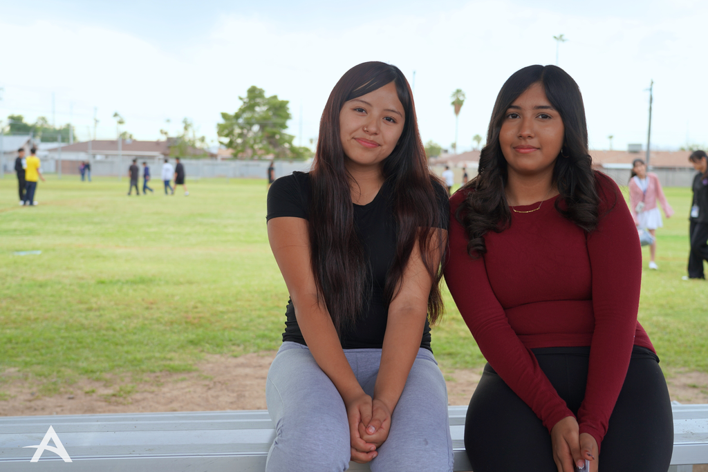 Two students sitting together on bleachers at a grassy field, facing the camera.
