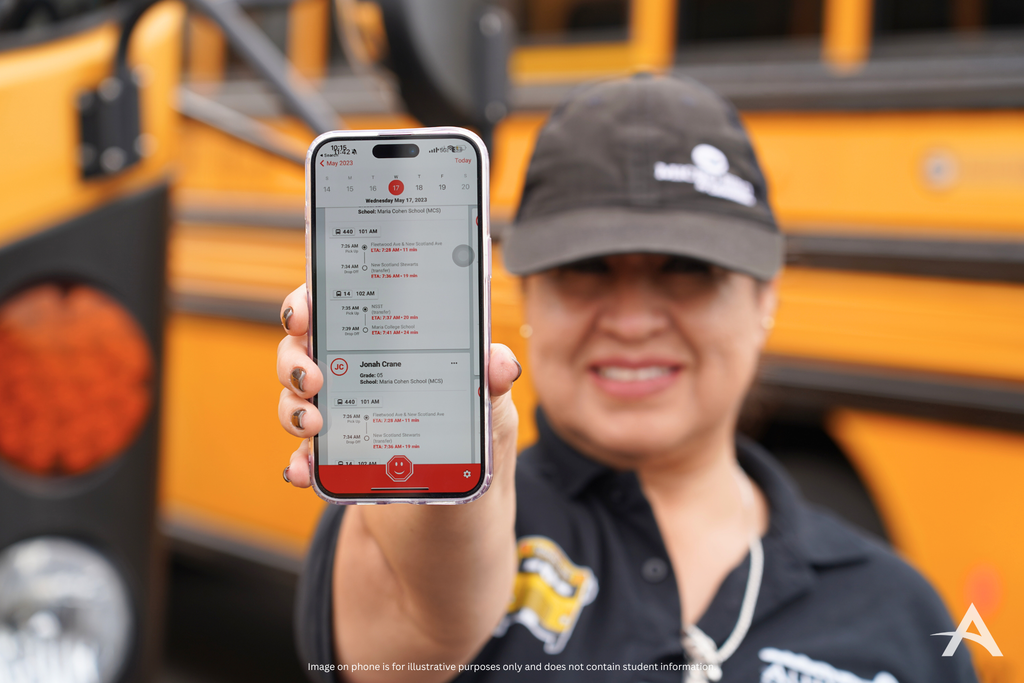 Person standing in front of a school bus holding a phone displaying the Stopfinder app with bus route and schedule information.