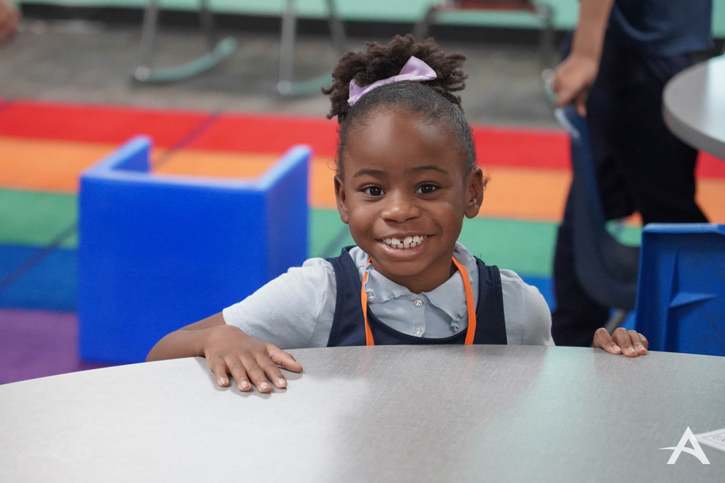 A smiling young student sitting at a classroom table, with colorful seating and a bright classroom environment in the background.