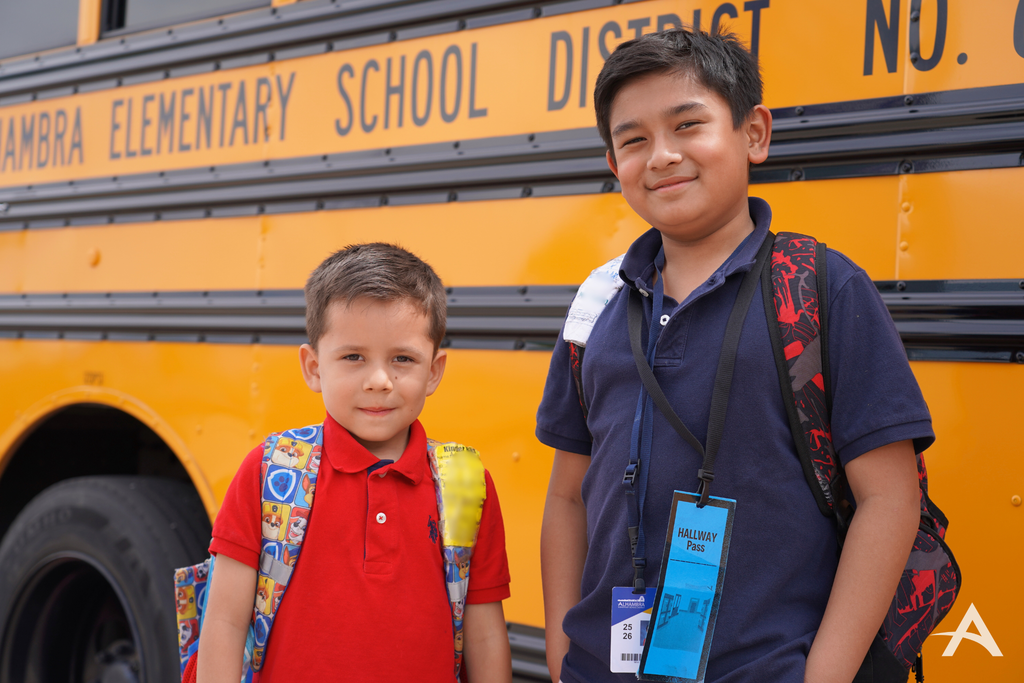 Two students with backpacks standing in front of a yellow school bus, smiling at the camera.