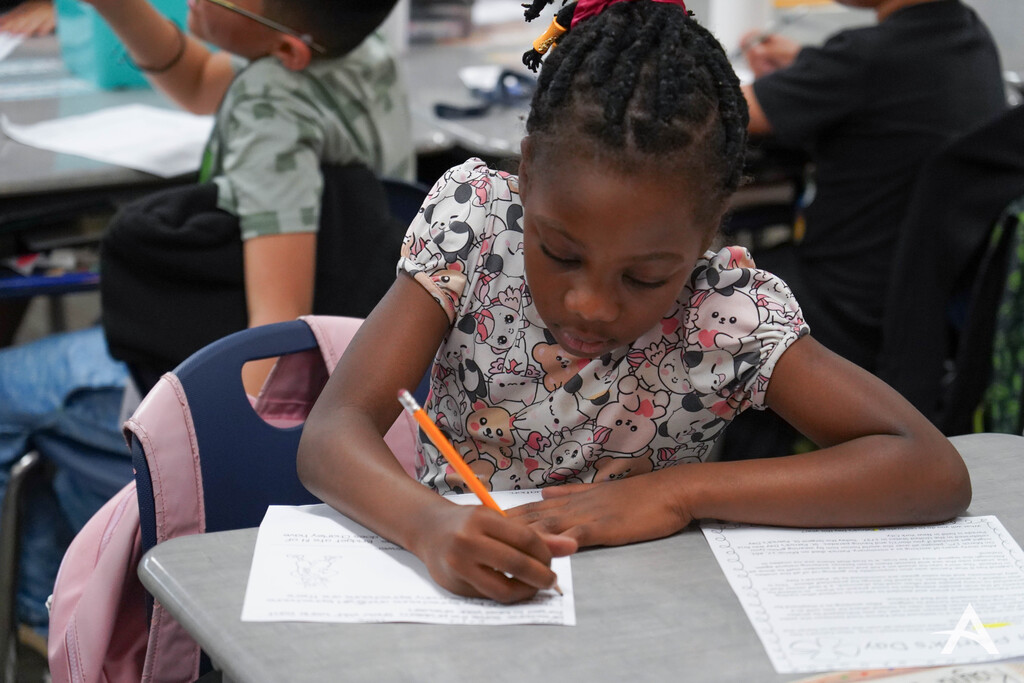 Image of a young girl at her desk with a pencil in hand working on school work. Some peers can be seen out of focus in the background.