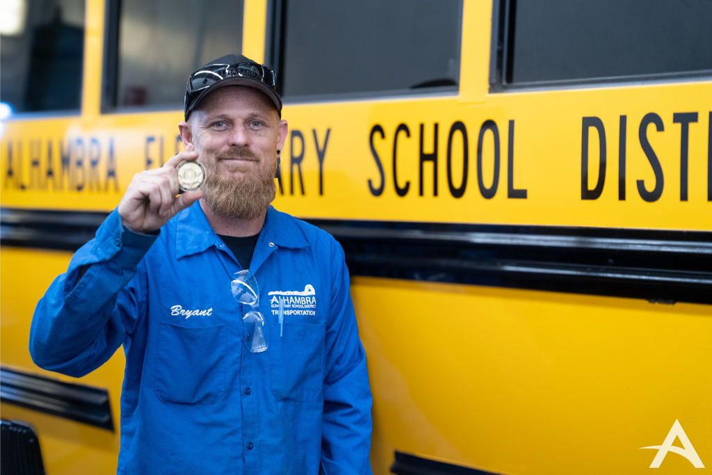Image of an Alhambra employee, Bryant, in front of a school bus holding up small medallion and smiling. 