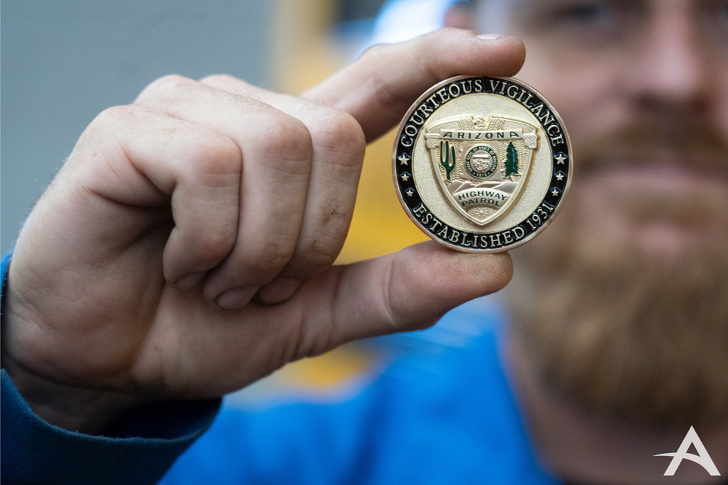 Close up image of the medallion between two fingers. The medallion reads, "Courteous Vigiliance, Established 1931," on the outer ring. Inner circle reads "Arizona Highway Patrol" and features a canyon, tree, and a cactus