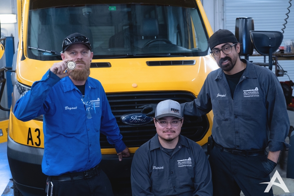 Image of 3 Alhambra employees in front of the front grill of a school bus. Left to right is Bryant, Patrick (who is sitting down), and Joseph. Bryant is holding a medallion.