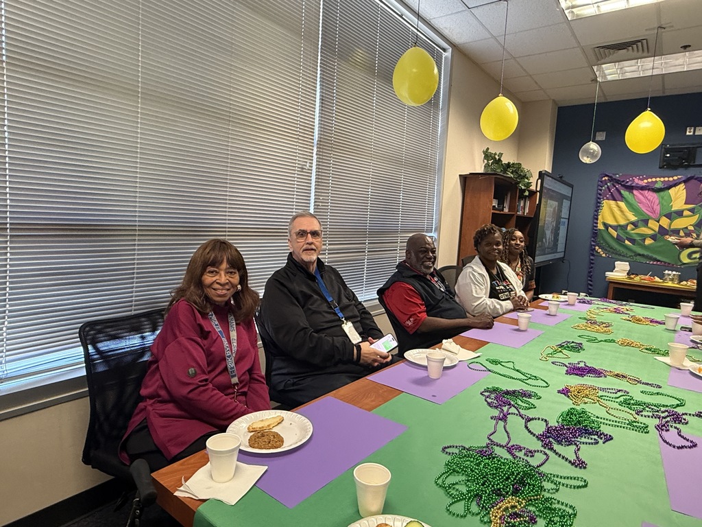 Five people sit at a table in a room with balloons and a banner. Four hold plates.