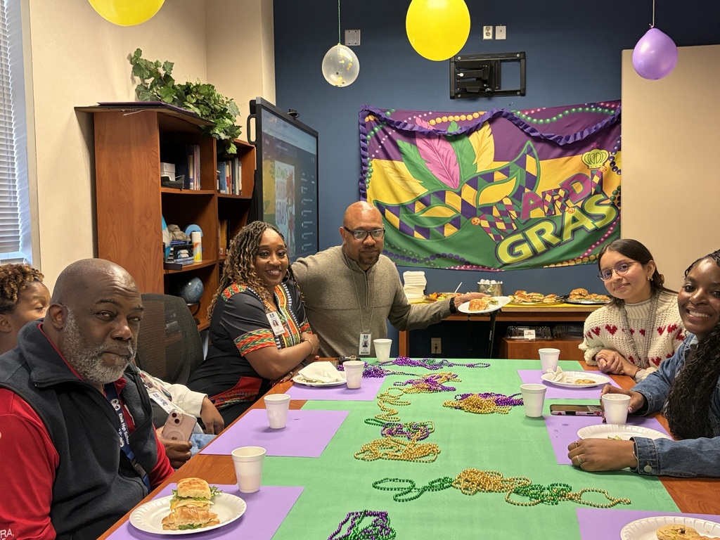 Group of people gathered around a table, some wearing festive beads, with a banner reading "GRAS" and balloons.