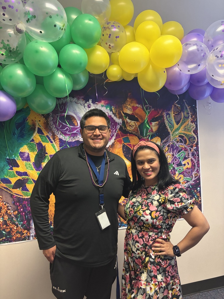 A man and woman stand next to each other, smiling. They are in front of a wall with balloons and a colorful painting.