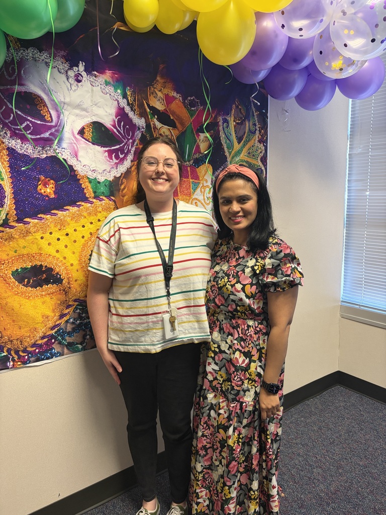 Two women stand in front of a wall with colorful balloons and a Mardi Gras mask backdrop.