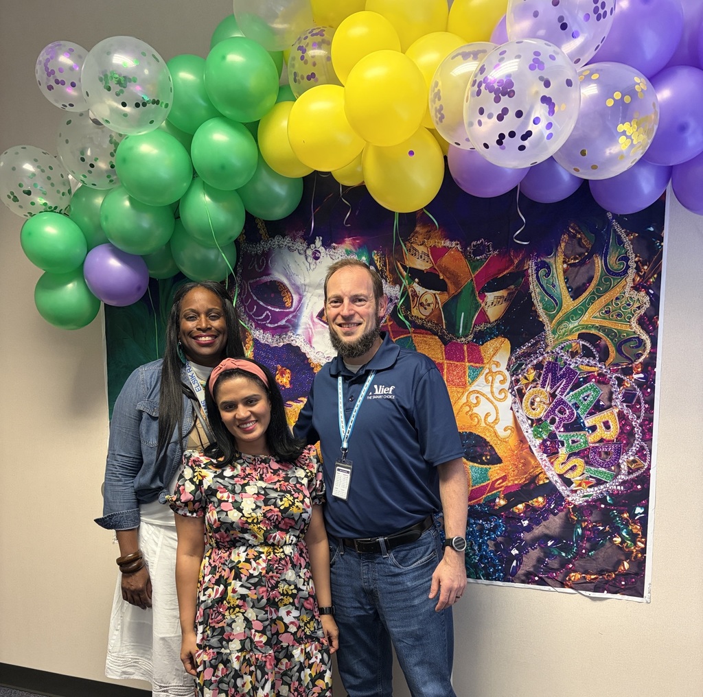 Three people stand near a colorful Mardi Gras banner. Two women and a man pose for a photo.