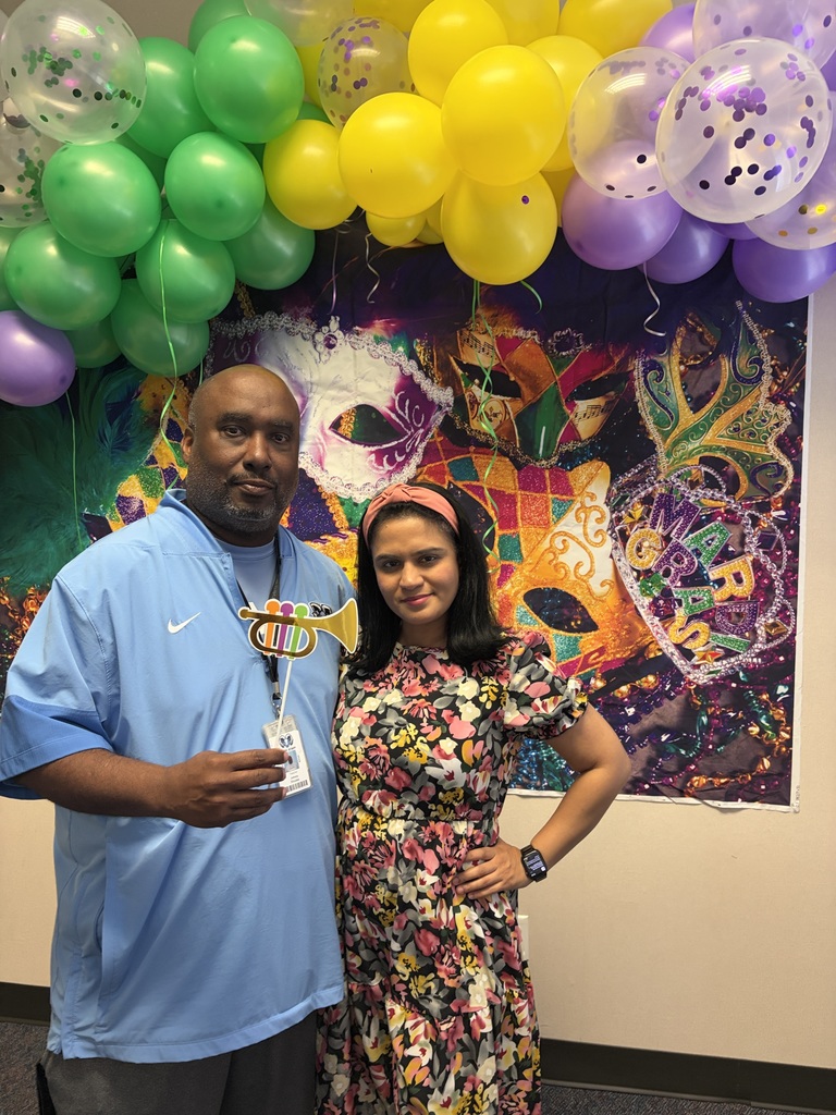 Man and woman stand next to a colorful Mardi Gras backdrop with balloons and a mask.