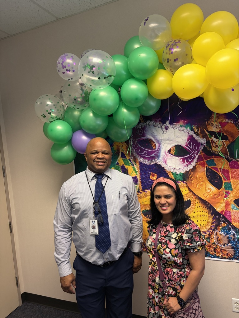 A man and woman stand next to each other in front of a wall decorated with colorful balloons and a Mardi Gras mask mural.