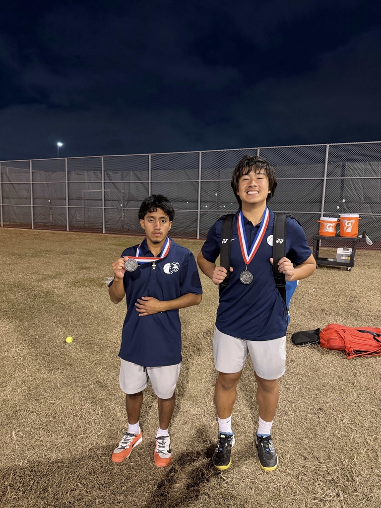 Two young men stand on a field, wearing blue shirts and white shorts. Both have medals around their necks.