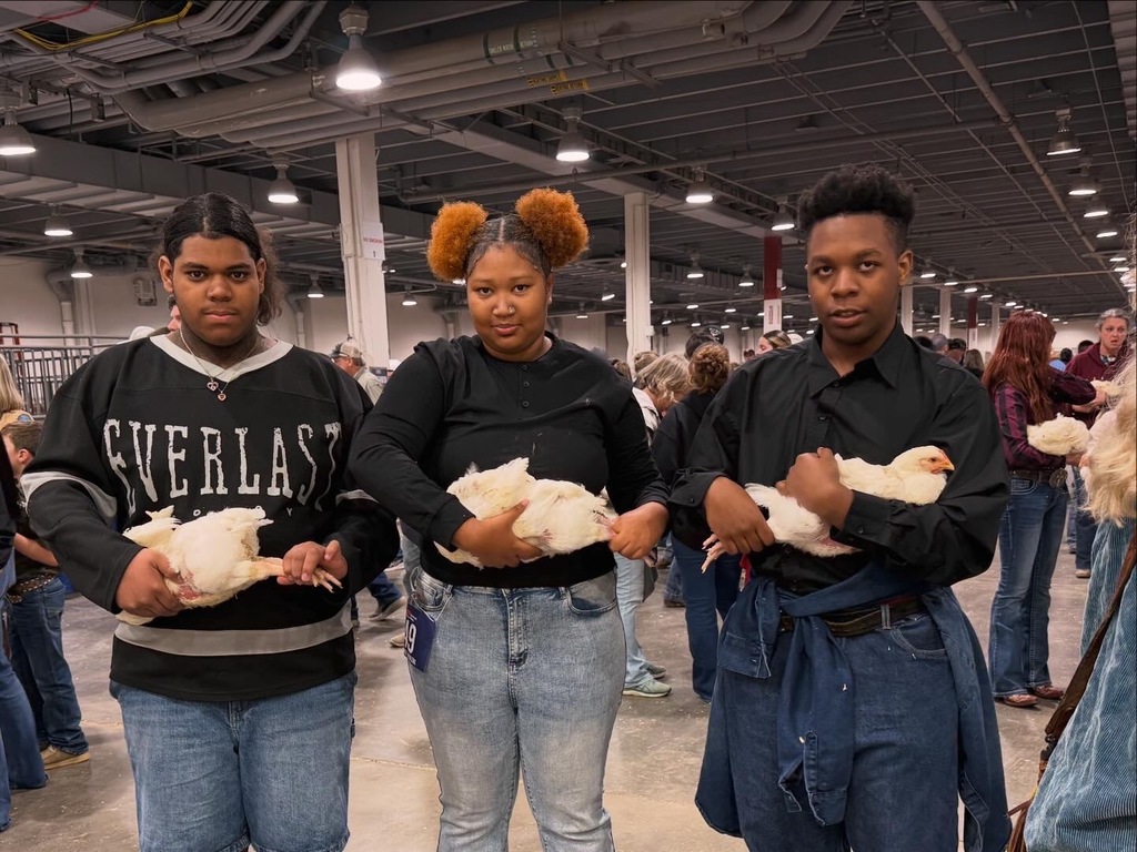 Three people stand in a large indoor area, each holding a white chicken. The ceiling is lit with overhead lights.