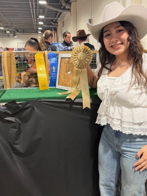 A woman in a white hat and blouse holds a golden ribbon while smiling. Behind her, people are near a display case with a dog inside.