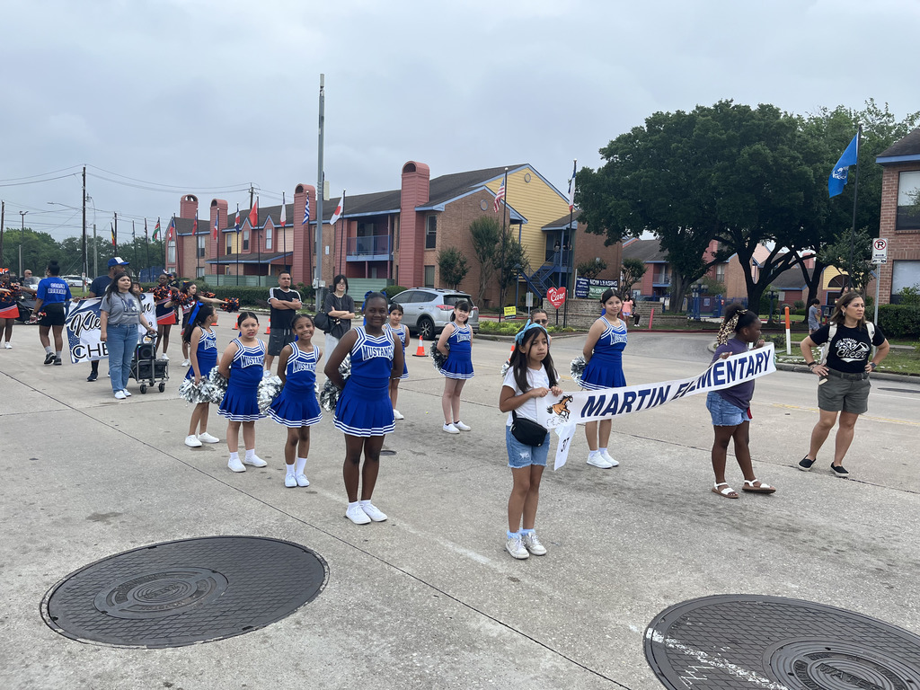 A group of young girls in blue uniforms and white shoes march in a parade. A banner reads "Martin Elementary".