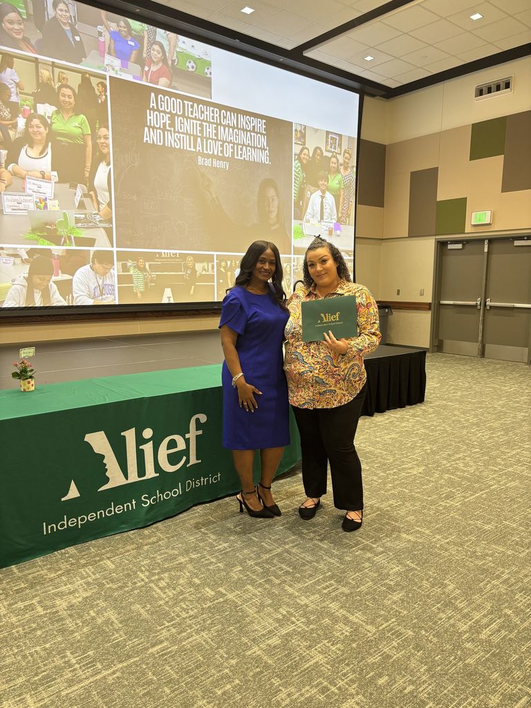 Two women, one holding a diploma, stand in front of a conference room screen and a banner.