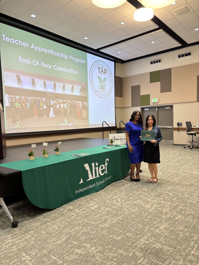 Two women in blue dresses stand in front of a table with a green tablecloth. A projector screen shows the text "Teacher Apprenticeship Program End-of-Year Celebration."