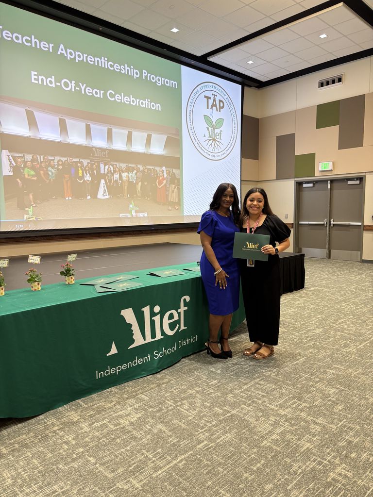 Two women stand by a table with a green tablecloth, holding certificates. A projector screen shows "Teacher Appreciation Program."