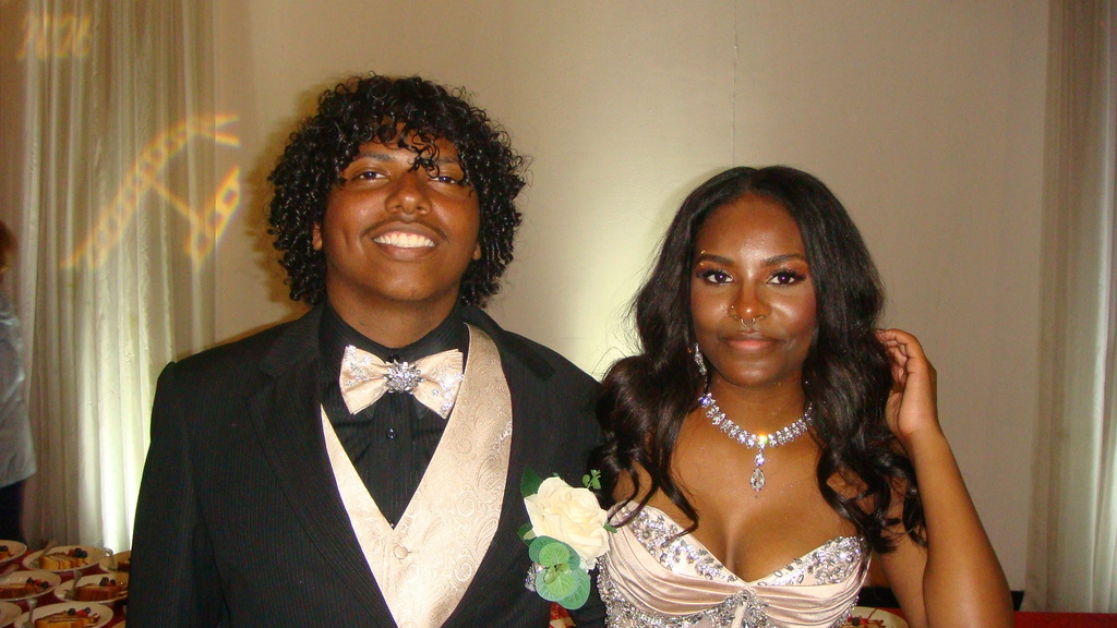 A man and woman in formal wear stand together, smiling. The man wears a black suit with a bow tie.
