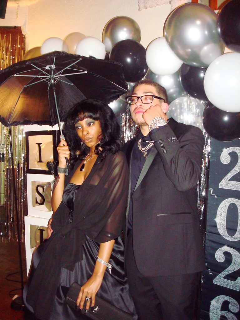 Couple in formal wear under a black umbrella, standing near balloons and a backdrop that reads 2026.