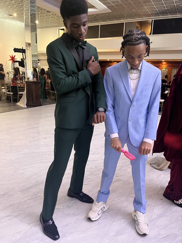 Two men, one in a green suit and the other in a blue suit, stand together indoors. The man in blue holds a pink object.