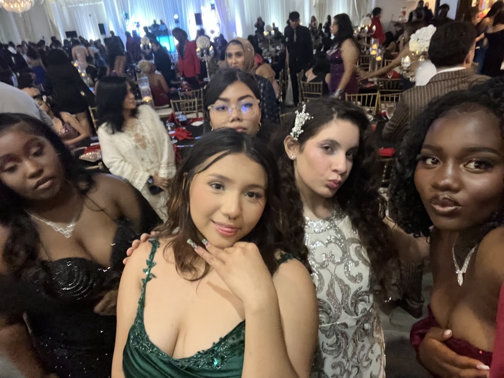 Five women in evening attire at an event, smiling and posing for a photo. They are standing close together with a large group of people behind them.