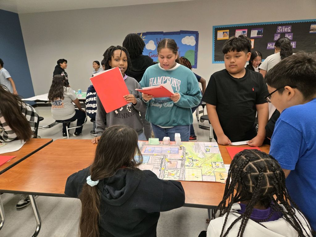 A group of students gather around a table with a map, discussing and looking at books.
