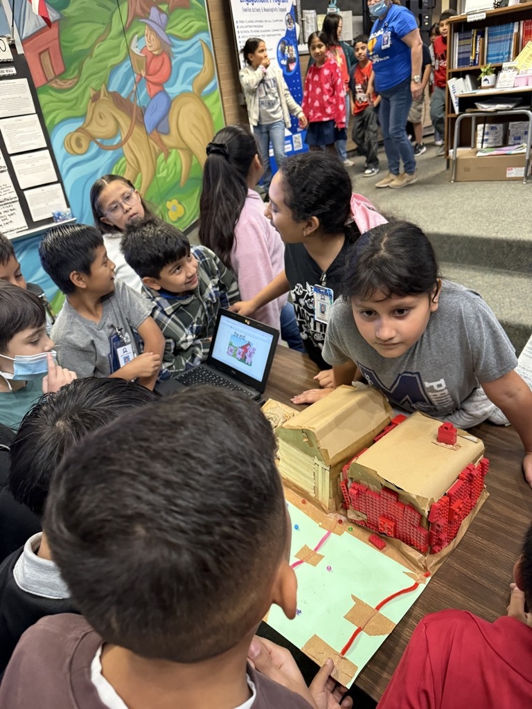 Children examine a cardboard house on a table in a classroom, some looking at a laptop.