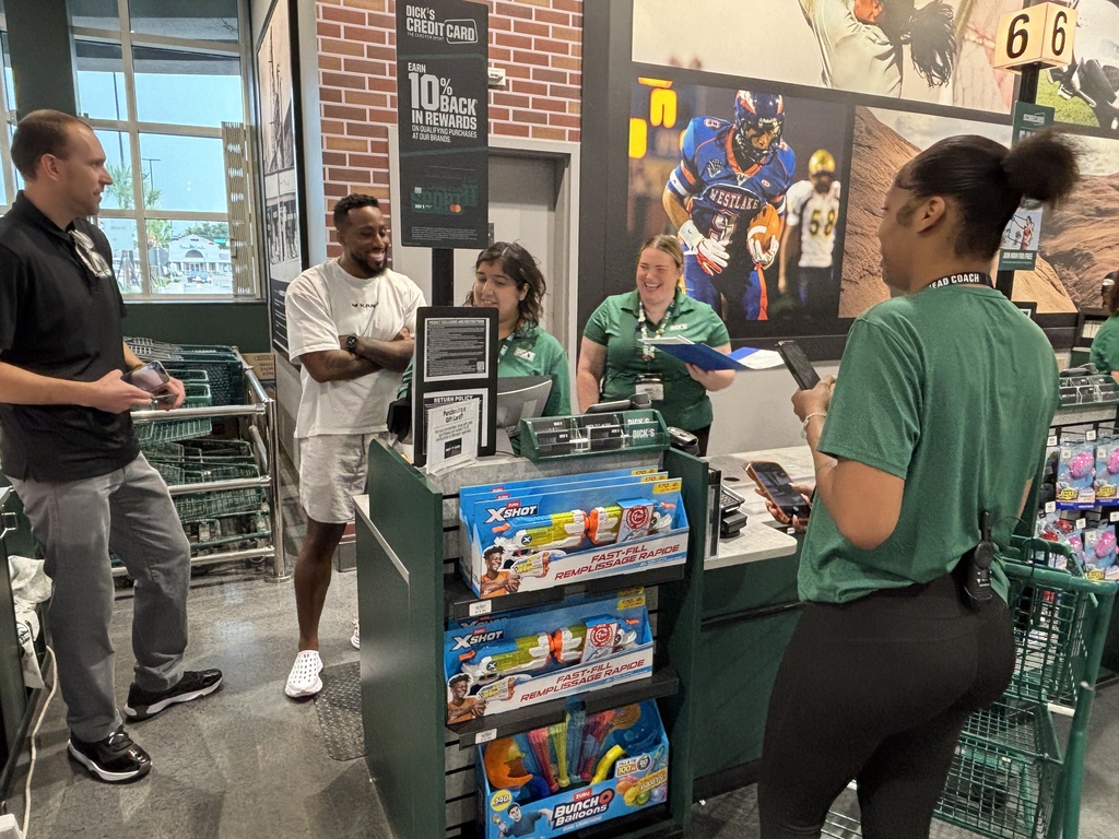 People in a store with shelves of toys and games. A woman in green interacts with a man in black.