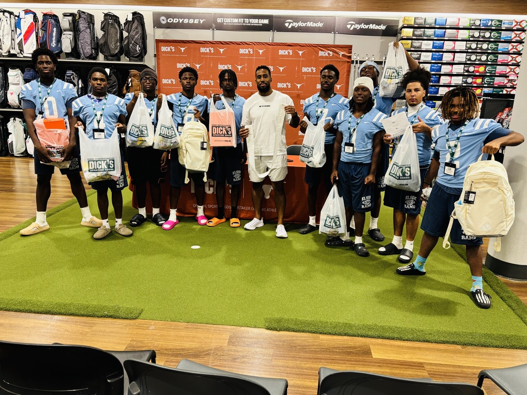 A group of young men wearing blue shirts stands in a sports store, holding bags, with a green carpet and chairs in the background.