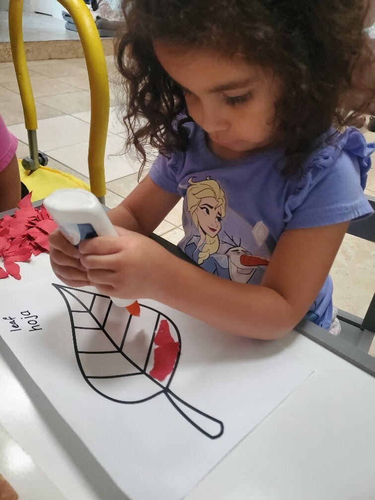 A child with curly hair uses a glue stick to apply red paint to a leaf drawing.