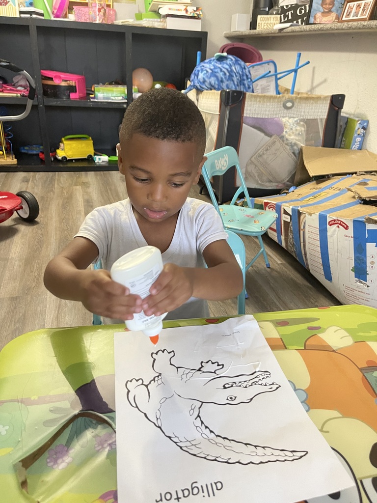 A child sits at a table applying glue to a coloring page featuring an alligator. Behind them is a playroom with shelves, chairs, and toys.
