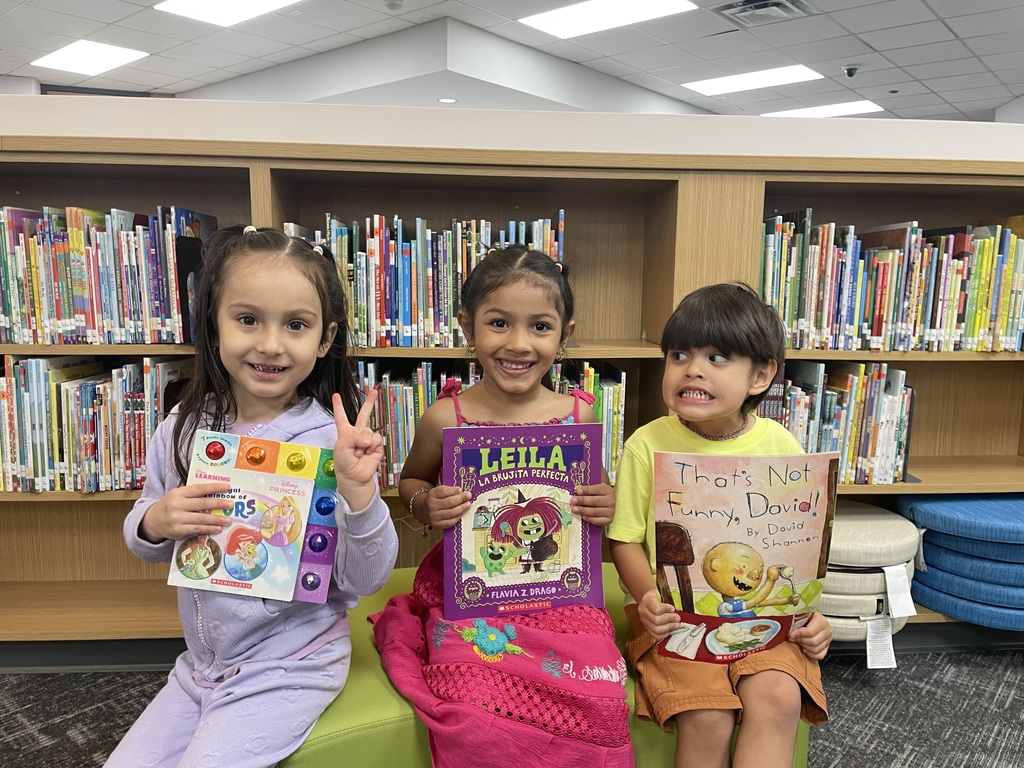 Three children seated in a library hold up books. Behind them are shelves filled with books and a blue cushion.