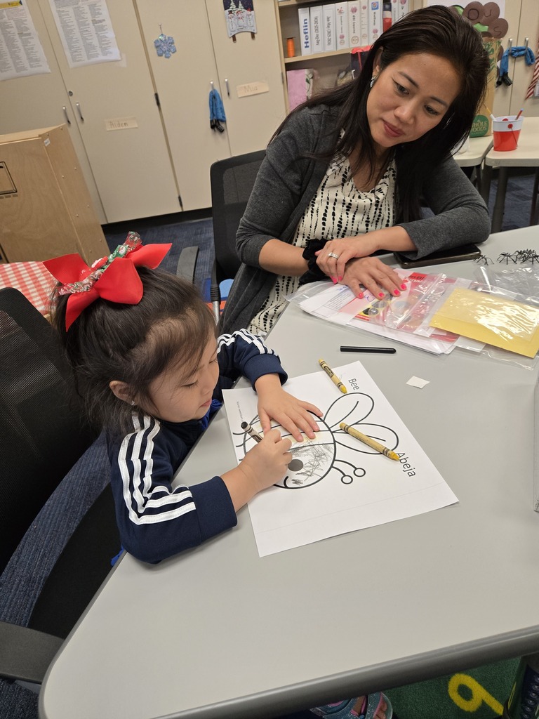 A young girl is seated at a table, drawing on a piece of paper with crayons. An adult woman sits next to her, offering assistance.