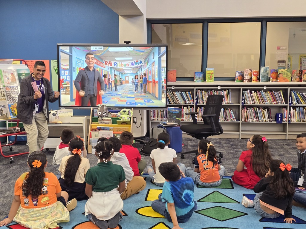 Mr. Garcia enthusiastically presents a Red Ribbon Week commercial on a large screen while Rees PVAA students watch from the library rug.