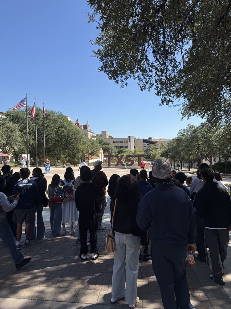 People stand on a sidewalk near a flagpole with trees and distant buildings under a clear sky.