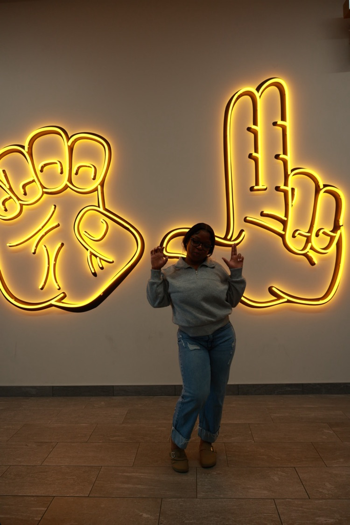 Person standing in front of neon hands on wall, making finger signs with both hands.